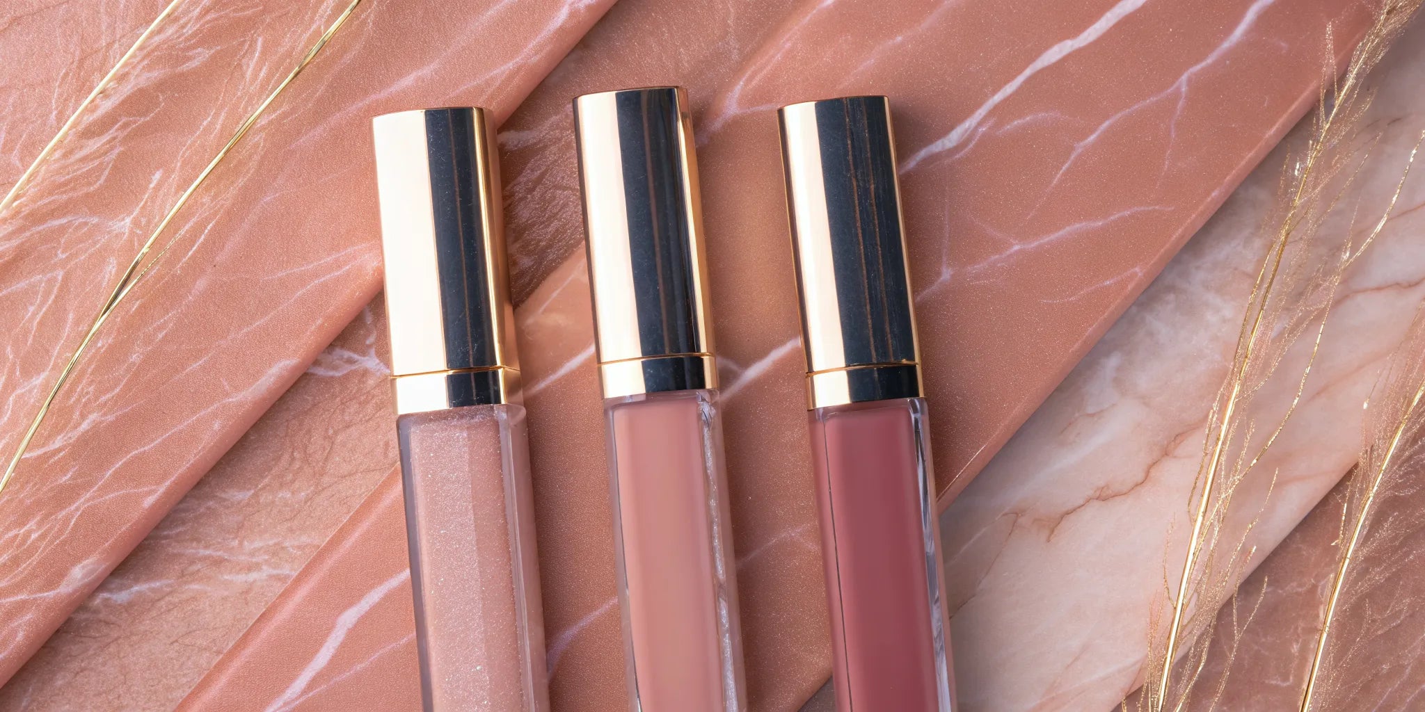 Three nude lip gloss shades in different tones arranged on marble surface with gold accents