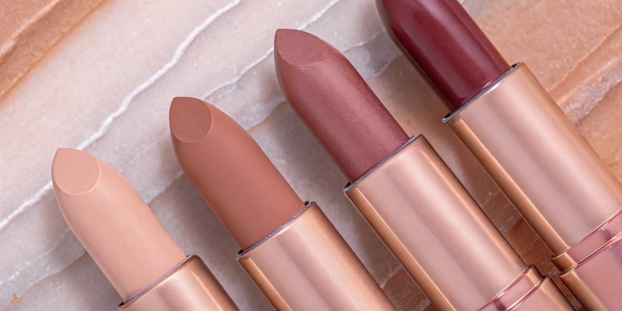 Four nude lipstick shades from light to dark arranged on marble with rose petals